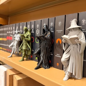 May include: A shelf displays several figurines of characters from The Lord of the Rings, each holding a book. The figurines are in various colors, including silver, green, and black, and are positioned in front of a row of books.