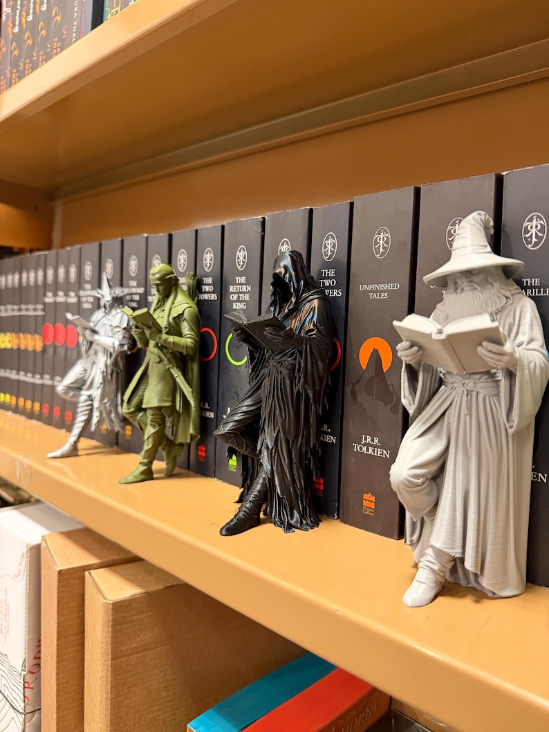 May include: A shelf displays several figurines of characters from The Lord of the Rings, each holding a book. The figurines are in various colors, including silver, green, and black, and are positioned in front of a row of books.
