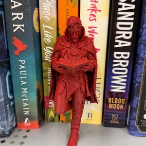 Puede incluir: Una figura roja de un personaje de cómic, de pie frente a una estantería de libros. La figura sostiene un libro y lleva una capa. Los libros tienen títulos como "Mistakes Were Made" y "Sandra Brown".