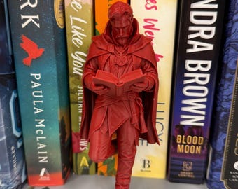 May include: A red figurine of a character from a comic book, standing in front of a shelf of books. The figurine is holding a book and wearing a cape. The books have titles like "Mistakes Were Made" and "Sandra Brown".