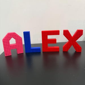 May include: The name "ALEX" constructed from colourful building blocks. The letters are pink, blue, red, and red. The blocks are on a black surface with a white background.