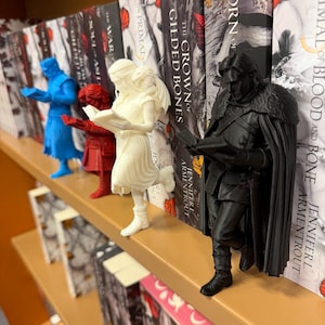 Puede incluir: Una estantería con varios sujetalibros de fantasía en azul, rojo, blanco y negro. Los sujetalibros son figuras de personas leyendo libros. Los libros tienen títulos como "Born of Blood and Bone" y "The Crown of Gilded Bones."