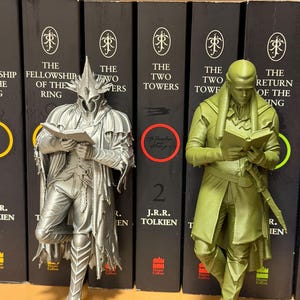 May include: Two figurines of characters from The Lord of the Rings, one silver and one green, are positioned in front of a row of books. The silver figure is of a character wearing a helmet and holding a book. The green figure is of a character holding a book.