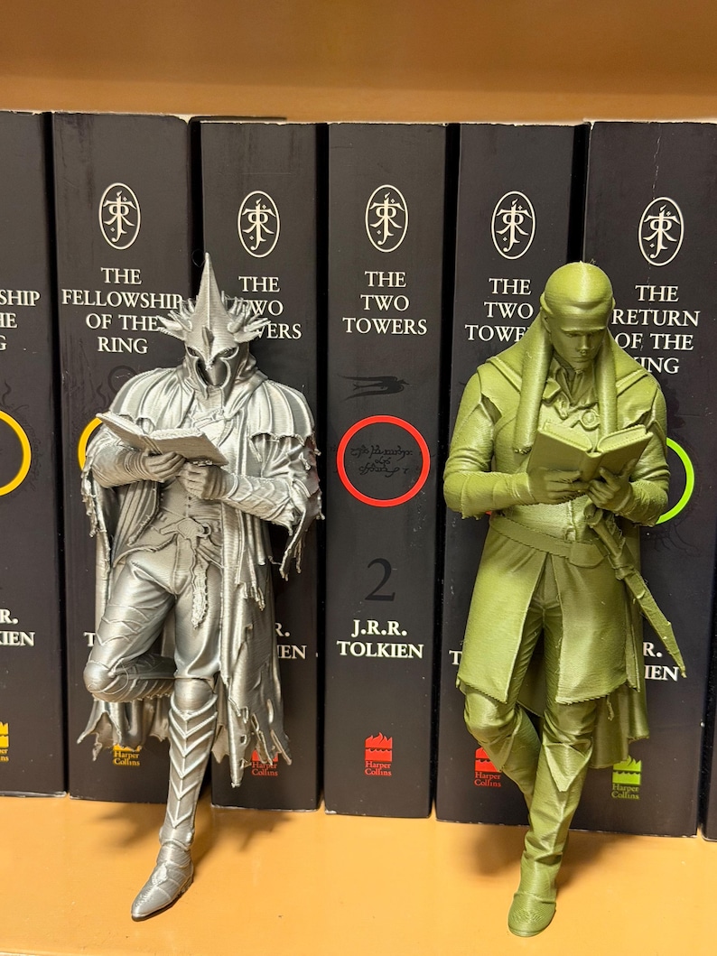 May include: Two figurines of characters from The Lord of the Rings, one silver and one green, are positioned in front of a row of books. The silver figure is of a character wearing a helmet and holding a book. The green figure is of a character holding a book.