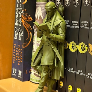 May include: A green figurine of an elf reading a book, leaning against a row of books. The figurine is detailed with a bow and quiver. The books include titles like "Emperor of Evening Stars" and "Beren and L&uacute;thien".