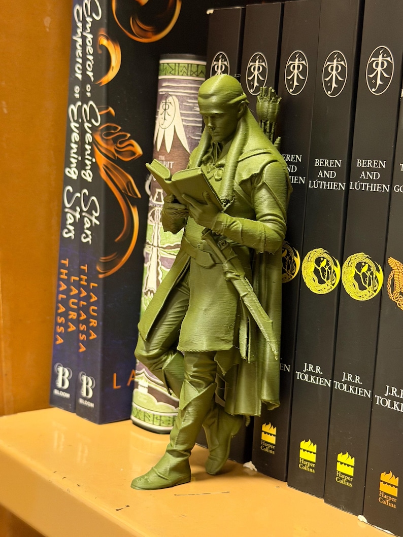 May include: A green figurine of an elf reading a book, leaning against a row of books. The figurine is detailed with a bow and quiver. The books include titles like "Emperor of Evening Stars" and "Beren and L&uacute;thien".
