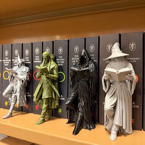 May include: Four figurines of characters from The Lord of the Rings, each holding a book, are displayed on a shelf. The figurines are in shades of gray, green, and black. Behind them are books with titles like "The Two Towers" and "The Return of the King."