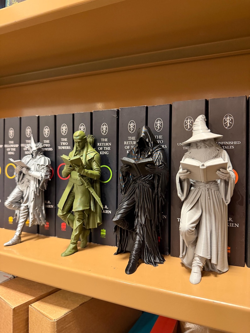 May include: Four figurines of characters from The Lord of the Rings, each holding a book, are displayed on a shelf. The figurines are in shades of gray, green, and black. Behind them are books with titles like "The Two Towers" and "The Return of the King."