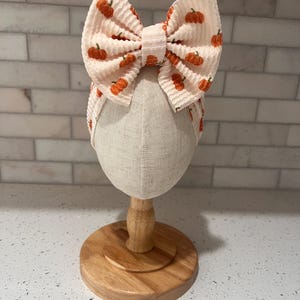 May include: A cream-colored fabric headband featuring a large, textured bow with a repeating orange pumpkin print. The headband is displayed on a neutral-toned mannequin head stand, showcasing the waffle-knit fabric texture and the pumpkin pattern.
