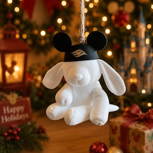 May include: A white plush dog ornament with long ears, wearing a black cap featuring the Disney Cruise Line logo. The toy is suspended by a piece of twine and has round black ears attached to the top of the cap, creating a character-inspired look.