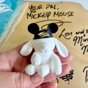 May include: A white figurine of a dog wearing a black Mickey Mouse hat with a gold emblem. The figurine is sitting down with its ears perked up. The background includes the text "Your Pal, Mickey Mouse" and other handwritten text.