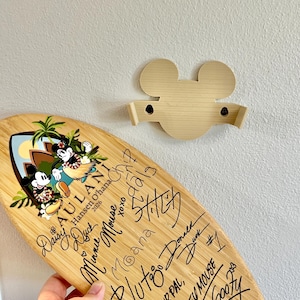 May include: A light-colored wooden surfboard-shaped plaque featuring a graphic of Mickey and Minnie Mouse in tropical attire with the text AULANI Hansen O'hana 2026. The plaque is covered in black ink signatures and a beige Mickey Mouse head wall mount.