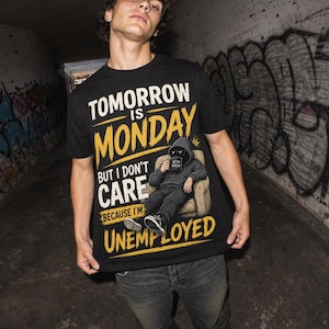 May include: A black t-shirt featuring a graphic of a person in a hoodie sitting in a chair holding a mug. The text reads: TOMORROW IS MONDAY BUT I DON'T CARE BECAUSE I'M UNEMPLOYED. The design uses yellow and white lettering on a black background.
