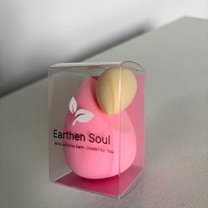 May include: A pink teardrop-shaped makeup sponge and a beige egg-shaped sponge are displayed in a clear plastic box. The box has the text "Earthen Soul" and "Handcrafted by Earth. Chosen by You."