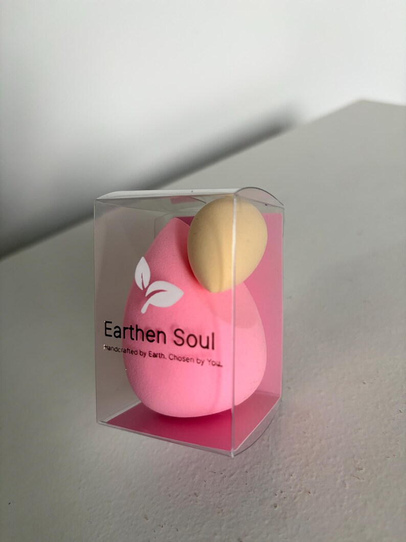 May include: A pink teardrop-shaped makeup sponge and a beige egg-shaped sponge are displayed in a clear plastic box. The box has the text "Earthen Soul" and "Handcrafted by Earth. Chosen by You."