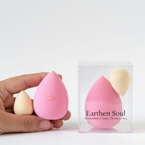May include: Pink and beige teardrop-shaped makeup sponges. One large pink sponge has a leaf design. The text "Earthen Soul" is on the clear box containing a pink sponge and a beige sponge. The sponges are for applying makeup.