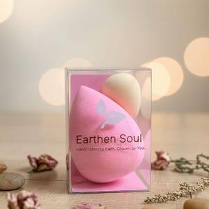 May include: A clear plastic box containing two makeup sponges. One is a large, pink teardrop shape, and the other is a smaller, beige egg shape. The pink sponge has the text "Earthen Soul" and "Handcrafted by Earth, Chosen by You."