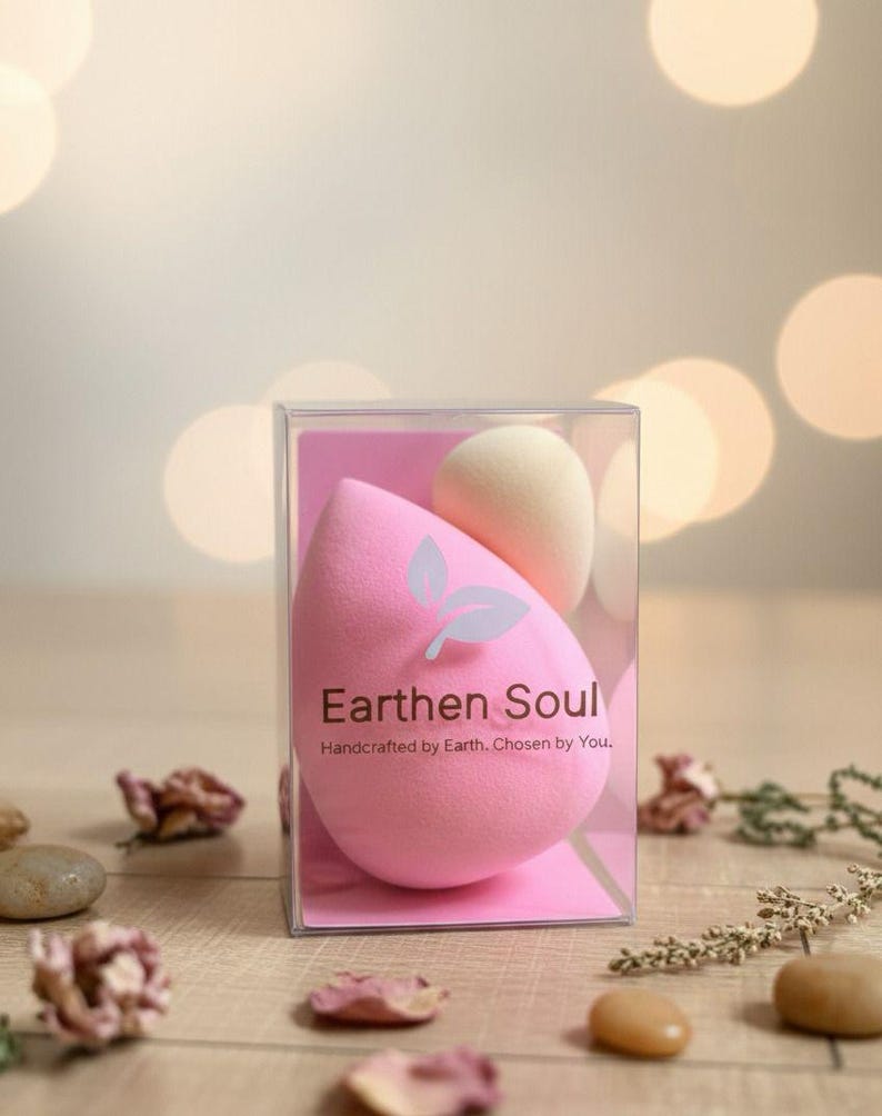 May include: A clear plastic box containing two makeup sponges. One is a large, pink teardrop shape, and the other is a smaller, beige egg shape. The pink sponge has the text "Earthen Soul" and "Handcrafted by Earth, Chosen by You."