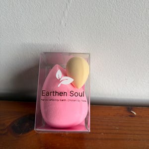 May include: A clear plastic box containing two makeup sponges. One is a large, pink teardrop shape, and the other is a smaller, yellow, rounded shape. The box has the text "Earthen Soul" and "Handcrafted by Earth. Chosen by You."