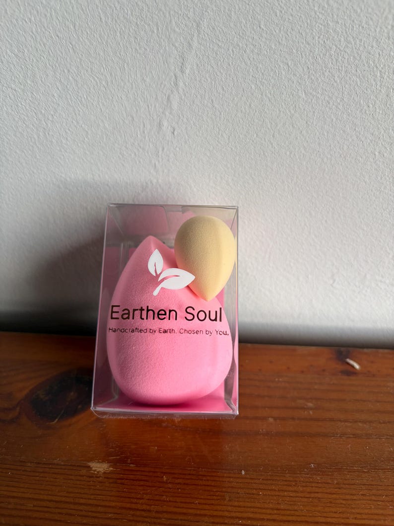 May include: A clear plastic box containing two makeup sponges. One is a large, pink teardrop shape, and the other is a smaller, yellow, rounded shape. The box has the text "Earthen Soul" and "Handcrafted by Earth. Chosen by You."
