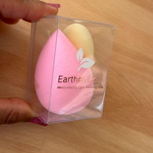 May include: A clear plastic box containing two makeup sponges. One is a light pink teardrop shape, and the other is a light yellow oval. The box has the text "Earthen Soul" and a leaf design.