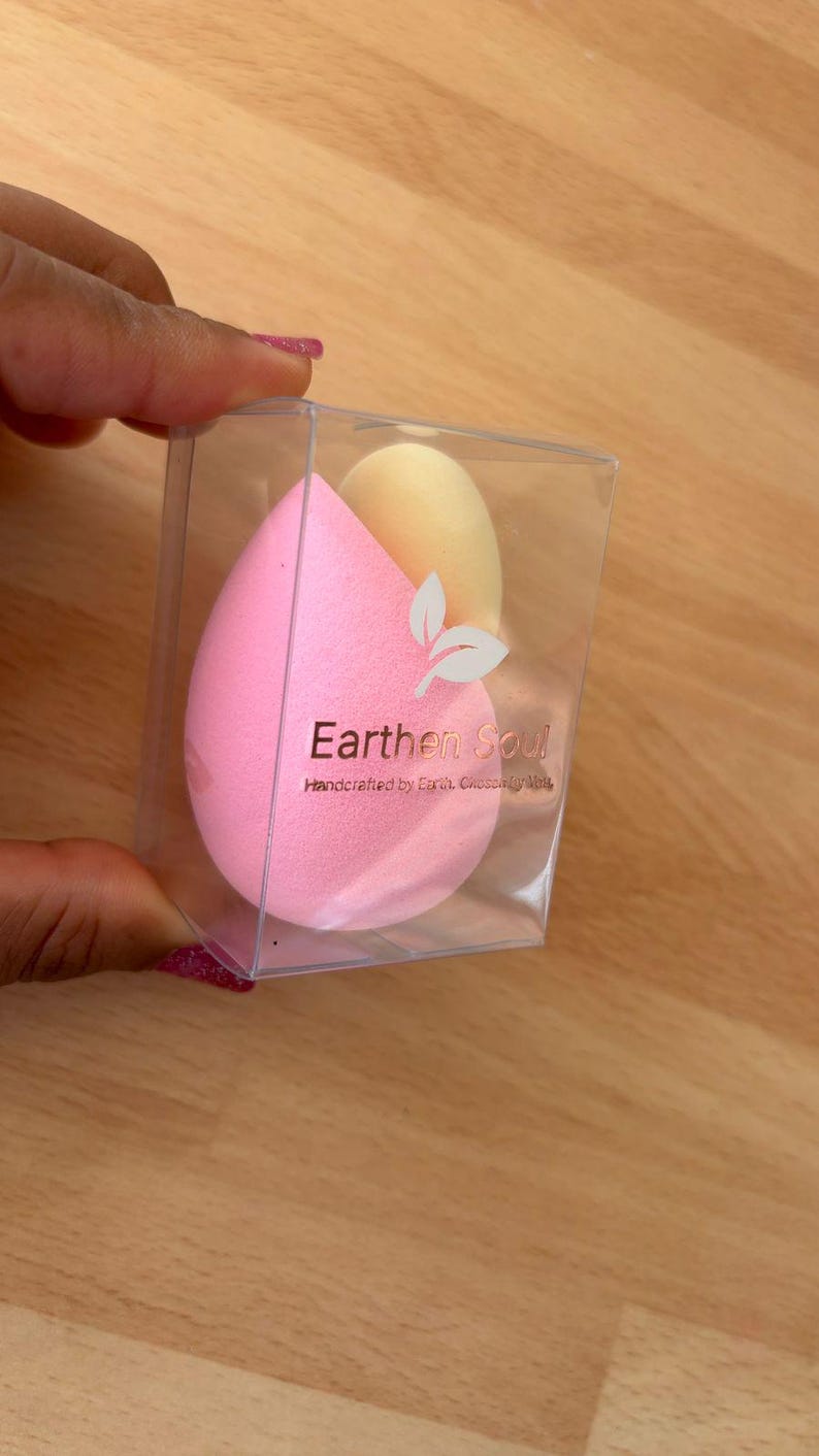 May include: A clear plastic box containing two makeup sponges. One is a light pink teardrop shape, and the other is a light yellow oval. The box has the text "Earthen Soul" and a leaf design.