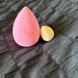 May include: Two makeup sponges. A large, pink, teardrop-shaped sponge with a leaf design and a smaller, yellow, egg-shaped sponge. Both are made of a soft, porous material. The sponges are on a dark, textured surface.