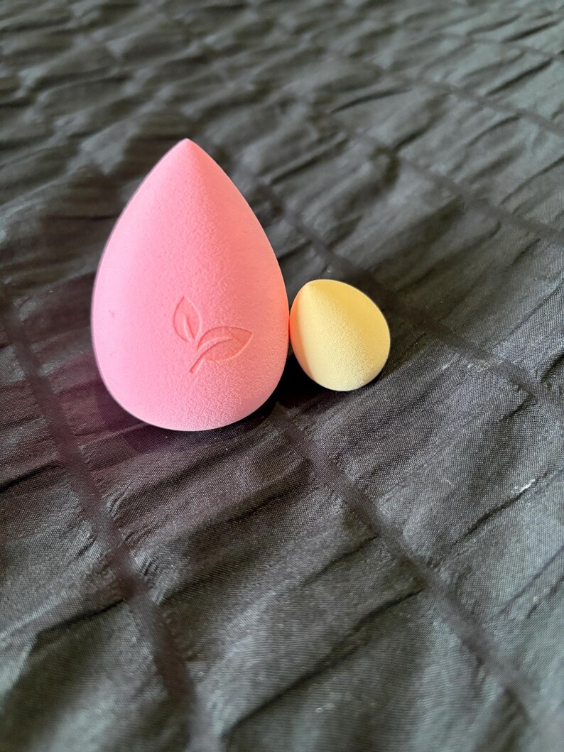 May include: Two makeup sponges. A large, pink, teardrop-shaped sponge with a leaf design and a smaller, yellow, egg-shaped sponge. Both are made of a soft, porous material. The sponges are on a dark, textured surface.