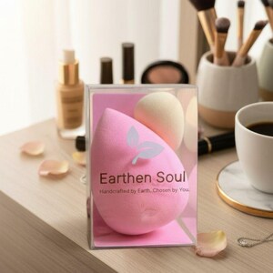 May include: A pink teardrop-shaped makeup sponge with a white leaf design and the text "Earthen Soul" in a clear plastic box. A beige egg-shaped sponge is also visible. Makeup brushes and a cup of coffee are in the background.