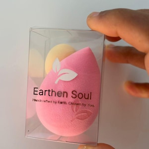 May include: A pink teardrop-shaped makeup sponge with a white leaf design and the text "Earthen Soul" in a clear plastic box. A smaller yellow sponge is also visible. The words "Handcrafted by Earth. Chosen by you." are below the main text.