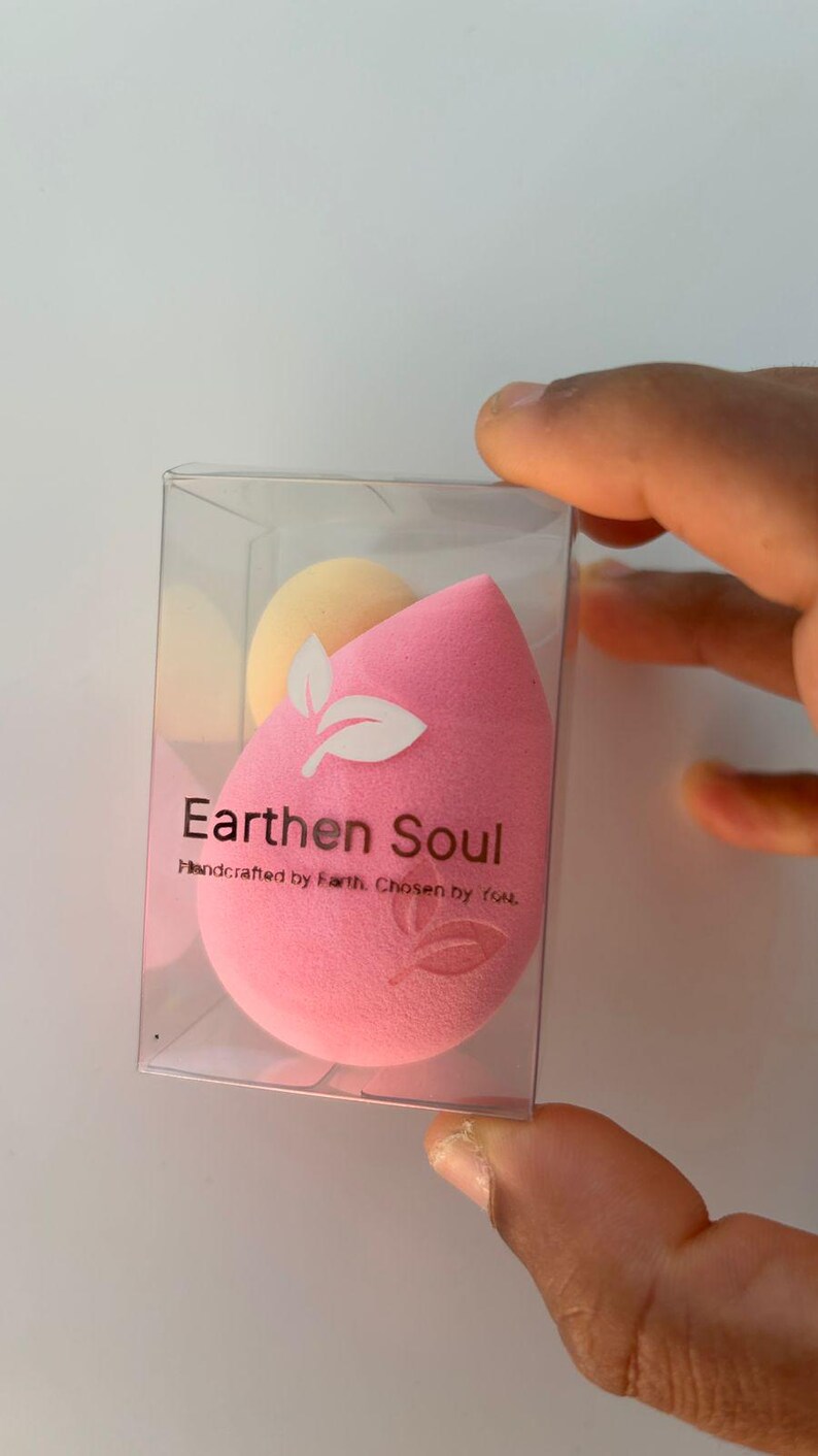 May include: A pink teardrop-shaped makeup sponge with a white leaf design and the text "Earthen Soul" in a clear plastic box. A smaller yellow sponge is also visible. The words "Handcrafted by Earth. Chosen by you." are below the main text.