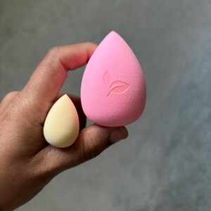 May include: Two teardrop-shaped makeup sponges. One is a large, bright pink with a leaf logo, and the other is a smaller, pale yellow. The sponges are held in a hand against a gray background. These are used for applying makeup.