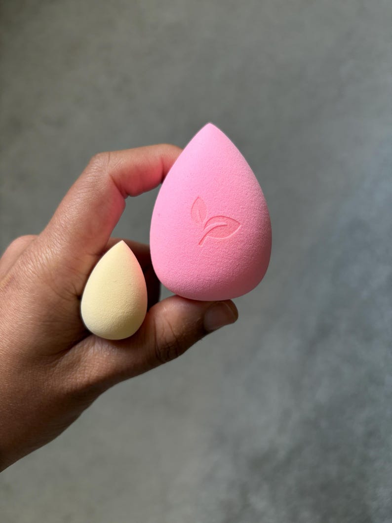 May include: Two teardrop-shaped makeup sponges. One is a large, bright pink with a leaf logo, and the other is a smaller, pale yellow. The sponges are held in a hand against a gray background. These are used for applying makeup.