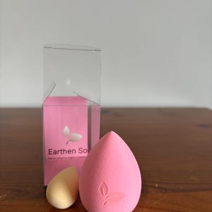 May include: A pink teardrop-shaped makeup sponge with a leaf design, next to a smaller yellow sponge. A pink box with the text "Earthen Soul" and a leaf logo is behind them. The sponges are on a wooden surface.