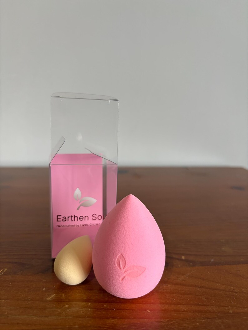 May include: A pink teardrop-shaped makeup sponge with a leaf design, next to a smaller yellow sponge. A pink box with the text "Earthen Soul" and a leaf logo is behind them. The sponges are on a wooden surface.