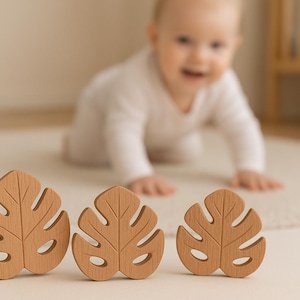 May include: Three wooden leaf-shaped toys with a light brown finish are arranged on a cream-colored surface. The toys are in focus, with a baby crawling in the background. The toys have a natural, organic aesthetic, suitable for baby play.