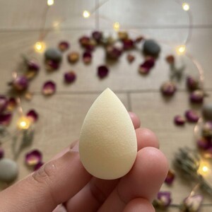 May include: A light beige teardrop-shaped makeup sponge. The sponge is held in a hand, with a blurred background of rose petals and fairy lights.