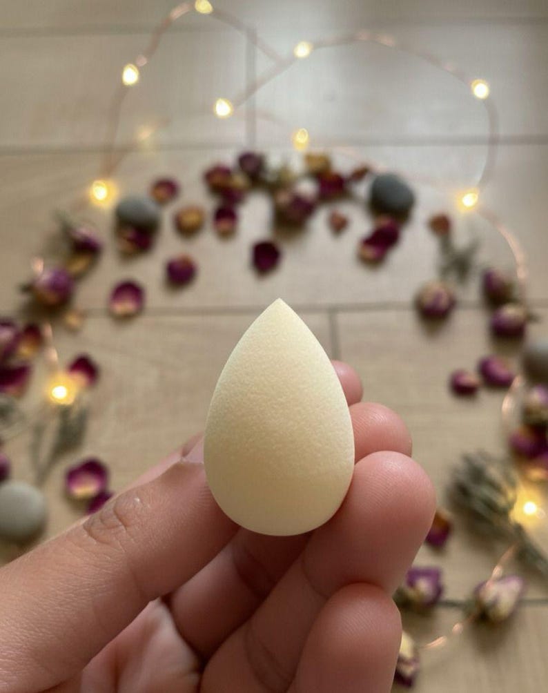 May include: A light beige teardrop-shaped makeup sponge. The sponge is held in a hand, with a blurred background of rose petals and fairy lights.