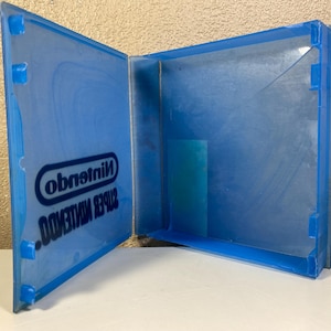 Puede incluir: Carcasa de juego Nintendo Super Nintendo azul transparente, abierta. El logotipo de Nintendo y el texto "SUPER NINTENDO" en blanco son visibles. La carcasa está hecha de un material plástico resistente. Para proteger el cartucho del juego.
