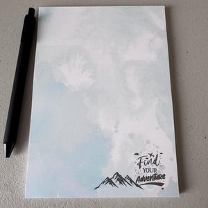 Peut inclure: Un bloc-notes rectangulaire avec un fond aquarelle bleu clair et blanc. Le coin inférieur droit présente un graphique noir de sommets montagneux et le texte "Find YOUR Adventure" avec de petites icônes d'avion et de nuages au-dessus.