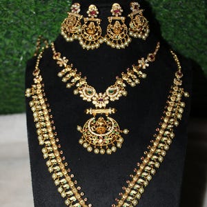 May include: Gold-toned jewelry set featuring a necklace, earrings, and a pendant. The necklace and earrings are adorned with pearls, gemstones, and intricate designs. The pendant showcases a detailed figure. The set is displayed on a black stand.