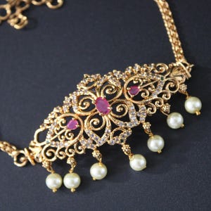 May include: Gold-toned necklace with an ornate, filigree pendant. The pendant features a central ruby-colored stone, smaller ruby accents, and clear crystals. White pearl drops hang from the bottom. The necklace has a delicate chain and is set against a dark background.