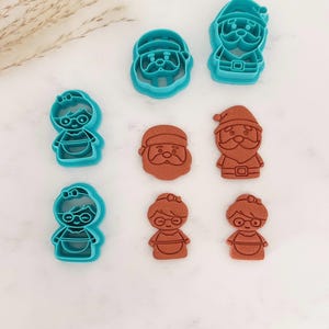 May include: A collection of four bright turquoise plastic cookie cutters and four corresponding brown stamped cookie impressions. The shapes include two figures with glasses and hair bows, a Santa Claus head, and a full-body Santa Claus figure.