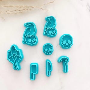 May include: A set of seven bright turquoise plastic cookie cutters featuring various shapes including two birds perched on skulls, a ghost, a small skull, a larger skull, a meat cleaver, a kitchen knife, and a curved scythe blade.