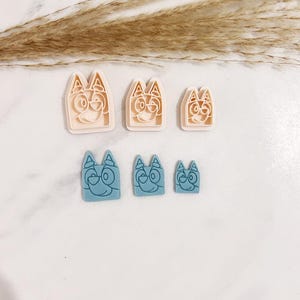 May include: A set of six cookie cutters in two colours and three sizes. The top row features three light peach cutters, and the bottom row features three blue cutters. Each cutter is shaped like a cartoon dog face with pointed ears and a snout.
