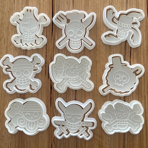 May include: Nine white plastic cookie cutters in various skull and crossbones designs. The cutters feature different hats, accessories, and expressions. The cutters are arranged on a wooden surface.