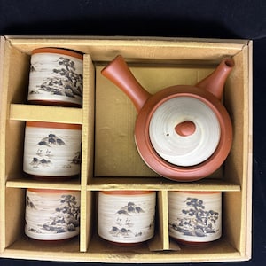 May include: A tea set in a cardboard box containing one terracotta teapot with a side handle and a light-colored lid, and five cylindrical tea cups. Each cup features a hand-painted black ink landscape design with trees and mountains.