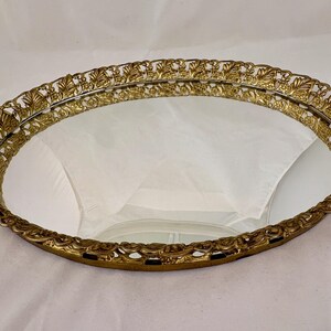 May include: An oval vanity tray featuring a reflective mirrored base and an ornate, gold-toned metal gallery rim. The raised border displays a repeating, intricate leaf-like filigree pattern that encircles the entire perimeter of the tray.