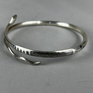 May include: Silver snake bracelet with detailed scales and a curved head and tail design. The bracelet wraps around, creating a unique and elegant accessory. The snake's body features a textured pattern.