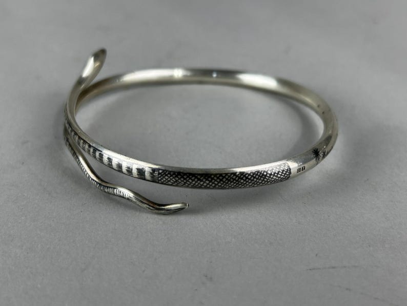 May include: Silver snake bracelet with detailed scales and a curved head and tail design. The bracelet wraps around, creating a unique and elegant accessory. The snake's body features a textured pattern.